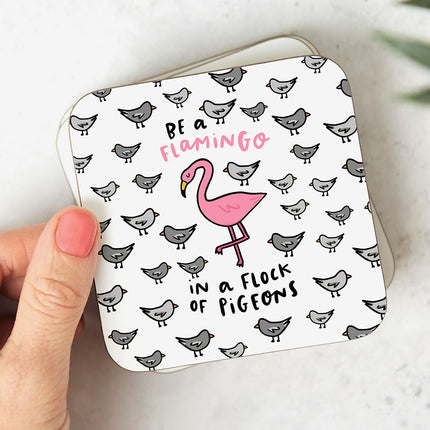 White coaster with a quirky illustration of a pink flamingo surrounded by pigeons. The text reads 'Be A Flamingo In A Flock Of Pigeons'.