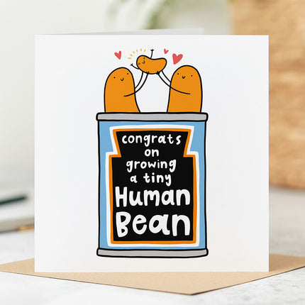 White greeting card with an illustration of a tin of beans with two beans at the top holding a baby bean. The Text reads 'Congrats On Growing  A Tiny Human Bean.
