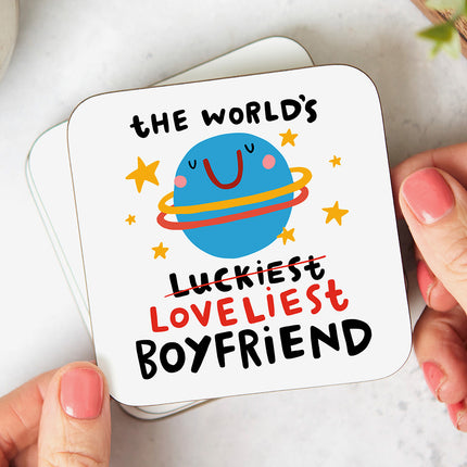 White coaster with a quirky planet illustration and the wording 'The World's Luckiest Loveliest Boyfriend' with the word luckiest crossed out.