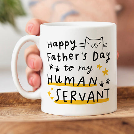 White ceramic mug that reads 'Happy Father's Day To My Human Servant' with a cat cartoon illustration and paws and stars.