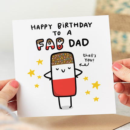 A white folded birthday card featuring a playful and colorful design with an image of a classic Fab ice lolly on the front. The card reads "Happy Birthday To A Fab Dad" in bold, eye-catching letters above the image. An arrow is pointing to the ice lolly, with the words "That's You" written next to it.