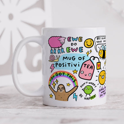 Image of a white ceramic mug with the words 'Mug of Positivi-Tea' written in playful lettering. The mug is decorated with various colourful illustrations and positive puns.